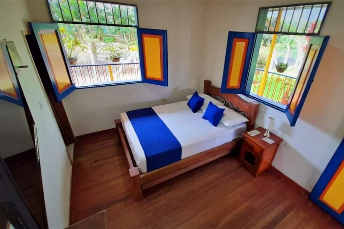 A bedroom with a wooden bed, blue and white bedding, a nightstand, and a window with a view of greenery.