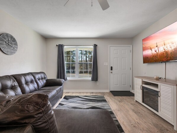 A living room with a leather sofa, a large window with curtains, a flat-screen TV on the wall, and a fireplace.