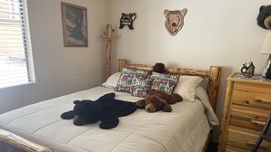 Een slaapkamer met een houten bedframe, een gewatteerde spreidek en twee kussens, evenals twee teddyberen. Er staat een houten dressoir met een klok en een klein beeldje erop.
