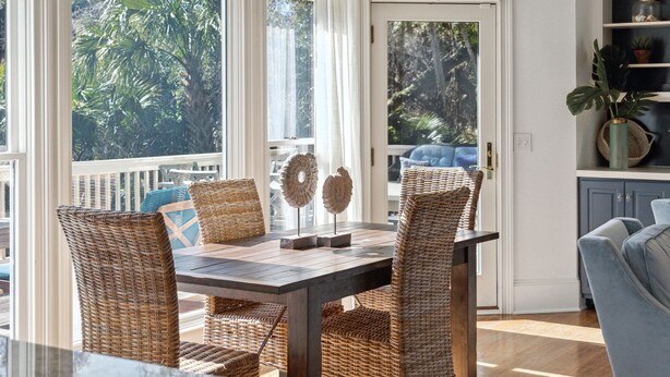 Een eethoek met rieten stoelen, een houten tafel en uitzicht op palmbomen door grote ramen.