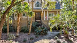 Een huis met twee verdiepingen, een veranda, een trap naar de ingang en een verzorgde tuin met palmbomen en struiken.