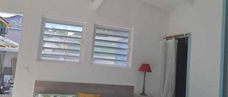Un dormitorio con cama, dos ventanas, una mesita de noche y una lámpara roja.
