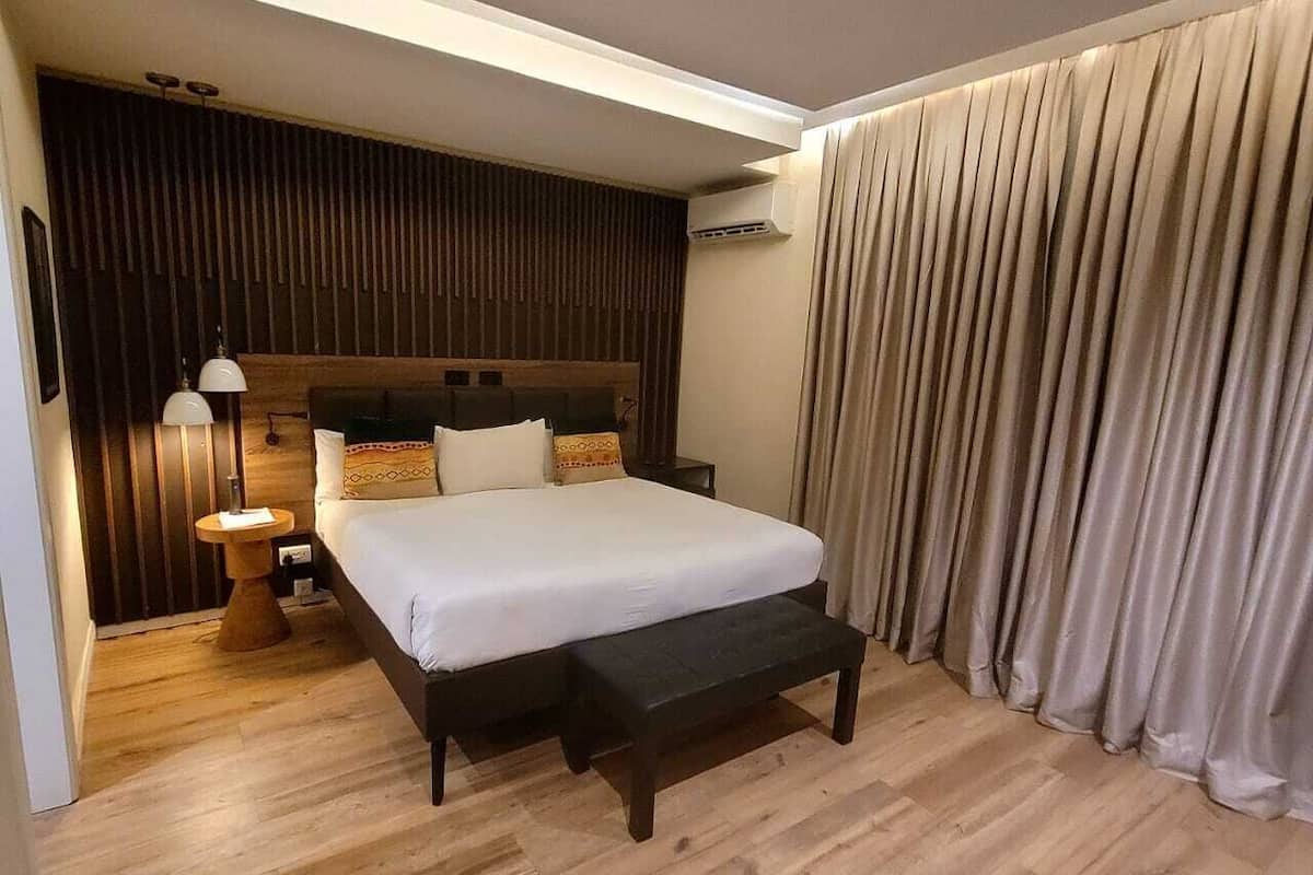 A modern hotel room with a large bed, wooden flooring, a headboard with vertical slats, a bedside table with a lamp, and a curtain on the window.