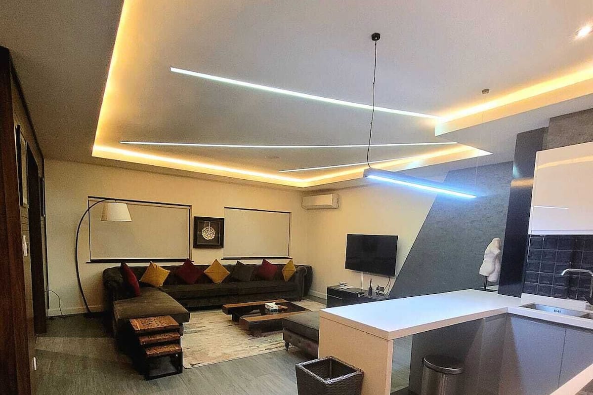 A modern living room with a sofa, coffee table, and TV. There is a kitchen area with a sink and a trash bin.