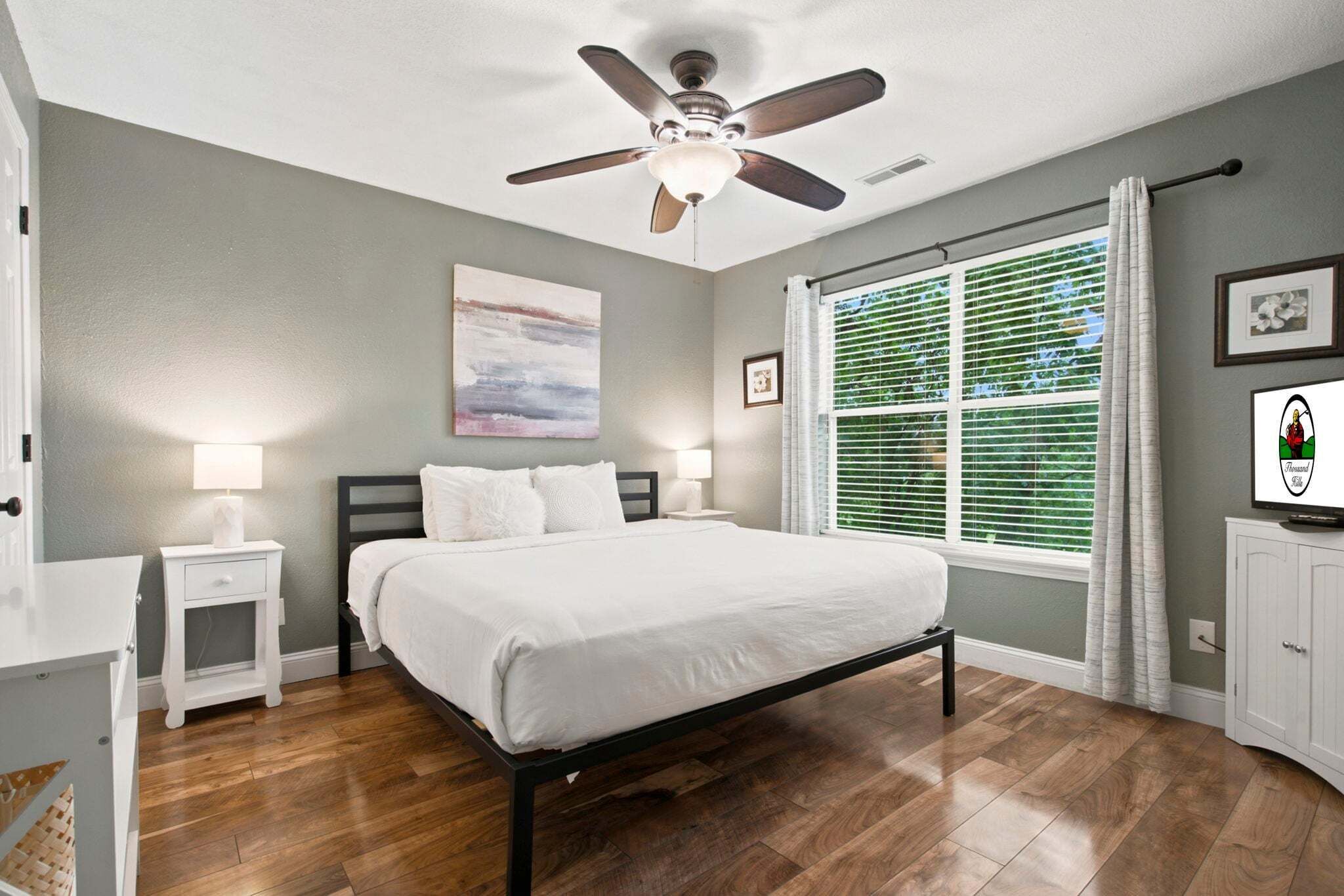 A bedroom with a bed, nightstands, a ceiling fan, a window with blinds, and a television.
