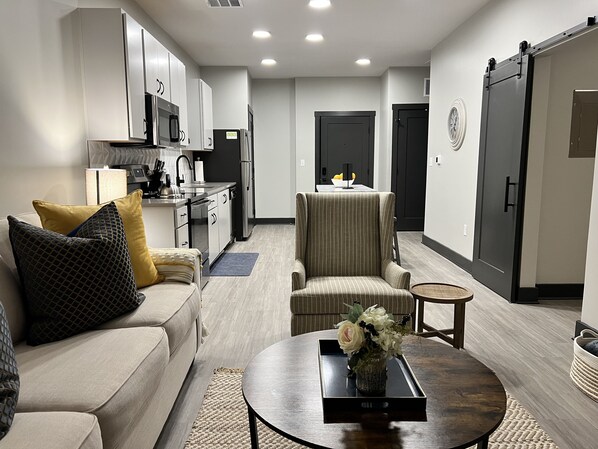 A modern living room with a sofa, armchair, coffee table, and a kitchen in the background.