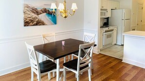 Un coin repas avec une table en bois foncé et des chaises blanches, un lustre et un tableau représentant un paysage accroché au mur.