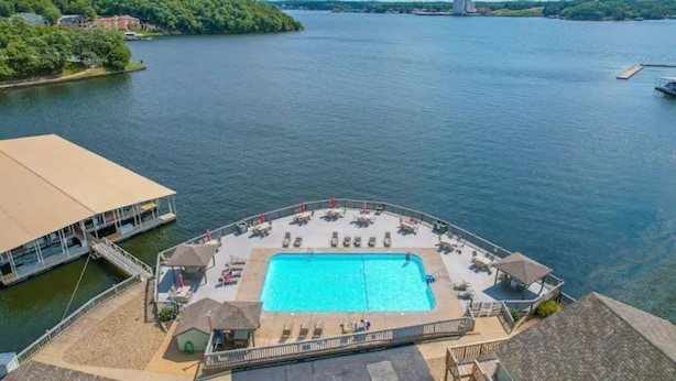 A large outdoor swimming pool surrounded by lounge chairs and umbrellas, with a view of a lake and distant buildings.