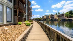 A riverside walkway with a wooden deck, a building with balconies, and a view of other buildings across the water.