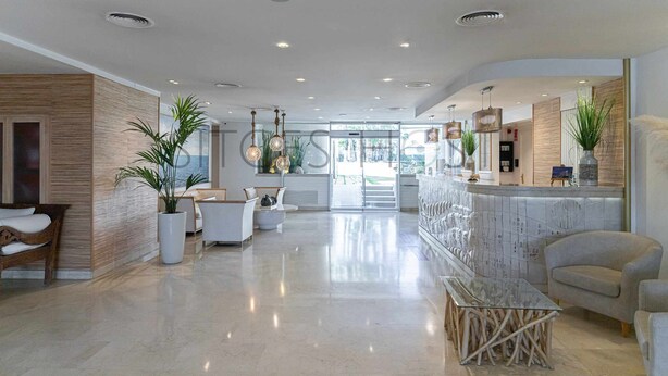 A spacious lobby with a reception desk, seating area, and a large window providing a view of the outdoors. - Sitges ARENA - Two pools by the beach (Sitges)