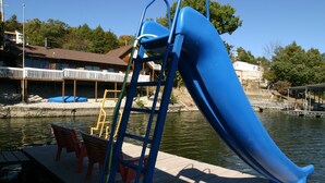Un parque infantil con un tobogán azul y una escalera amarilla, cerca de una masa de agua con un muelle y sillas.