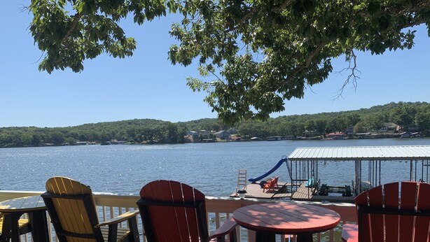 Terraza junto al lago con sillas Adirondack y una mesa, con vistas a un lago tranquilo y una costa lejana con casas.