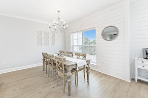 A dining room with a wooden table, chairs, a chandelier, and a large window with blinds. - Summer Salt Escape | Zero Entry Pool | Lake for Fishing | My Beach Getaways (Orange Beach)