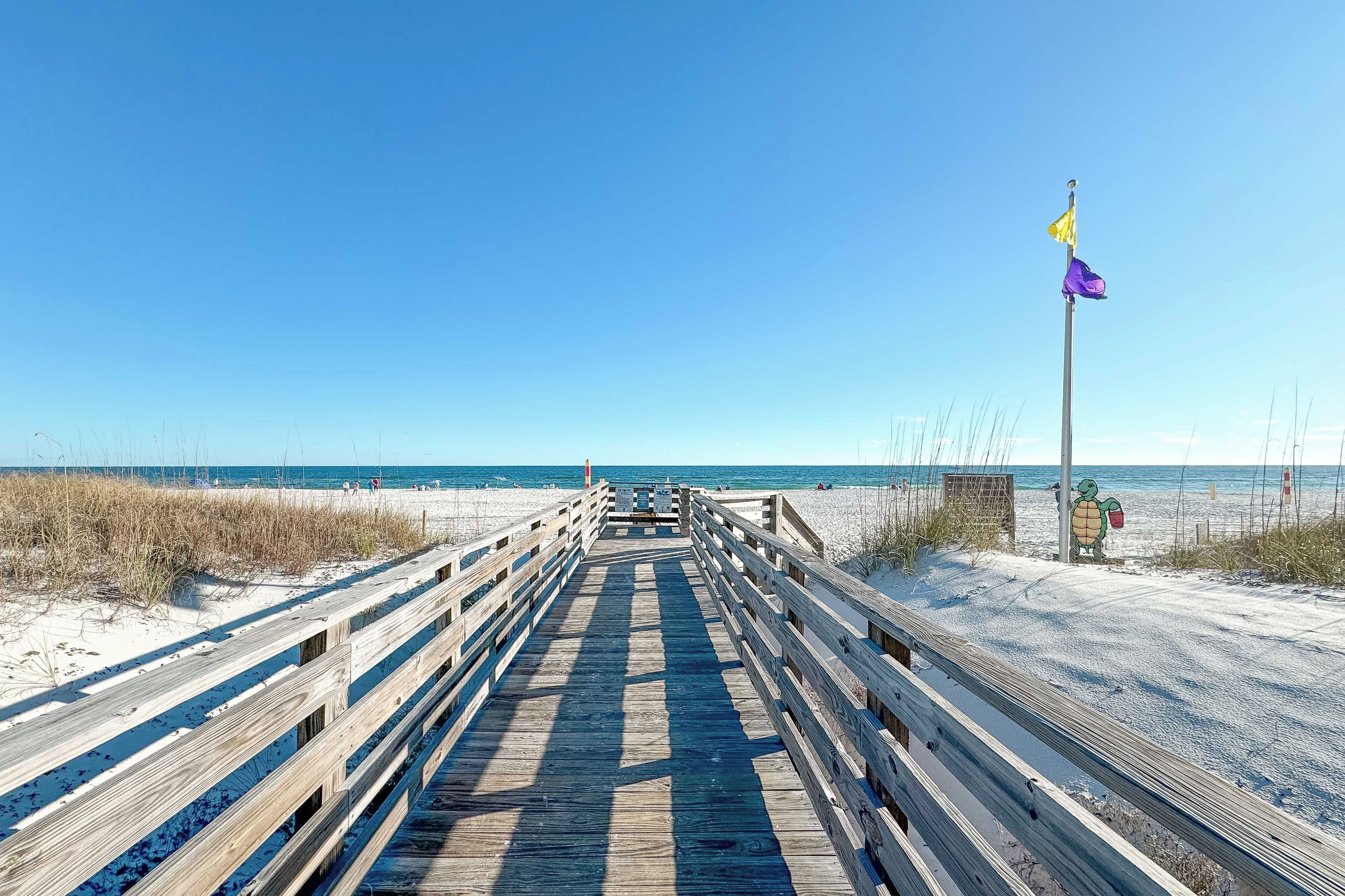A wooden boardwalk leading to a beach with a turtle statue and flags.