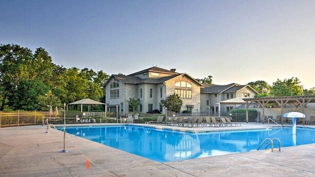 A large swimming pool with lounge chairs and umbrellas, surrounded by a stone building with multiple windows and a covered patio area. - Lissa's Retreat- 4 BDRM 3 BA Sleeps 12! Indoor Pool, Gym, Fishing, Golf, & Park (Branson)