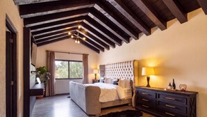 Une chambre à coucher comprenant un lit, des tables de chevet, une commode, une fenêtre et un plafond avec des poutres apparentes.