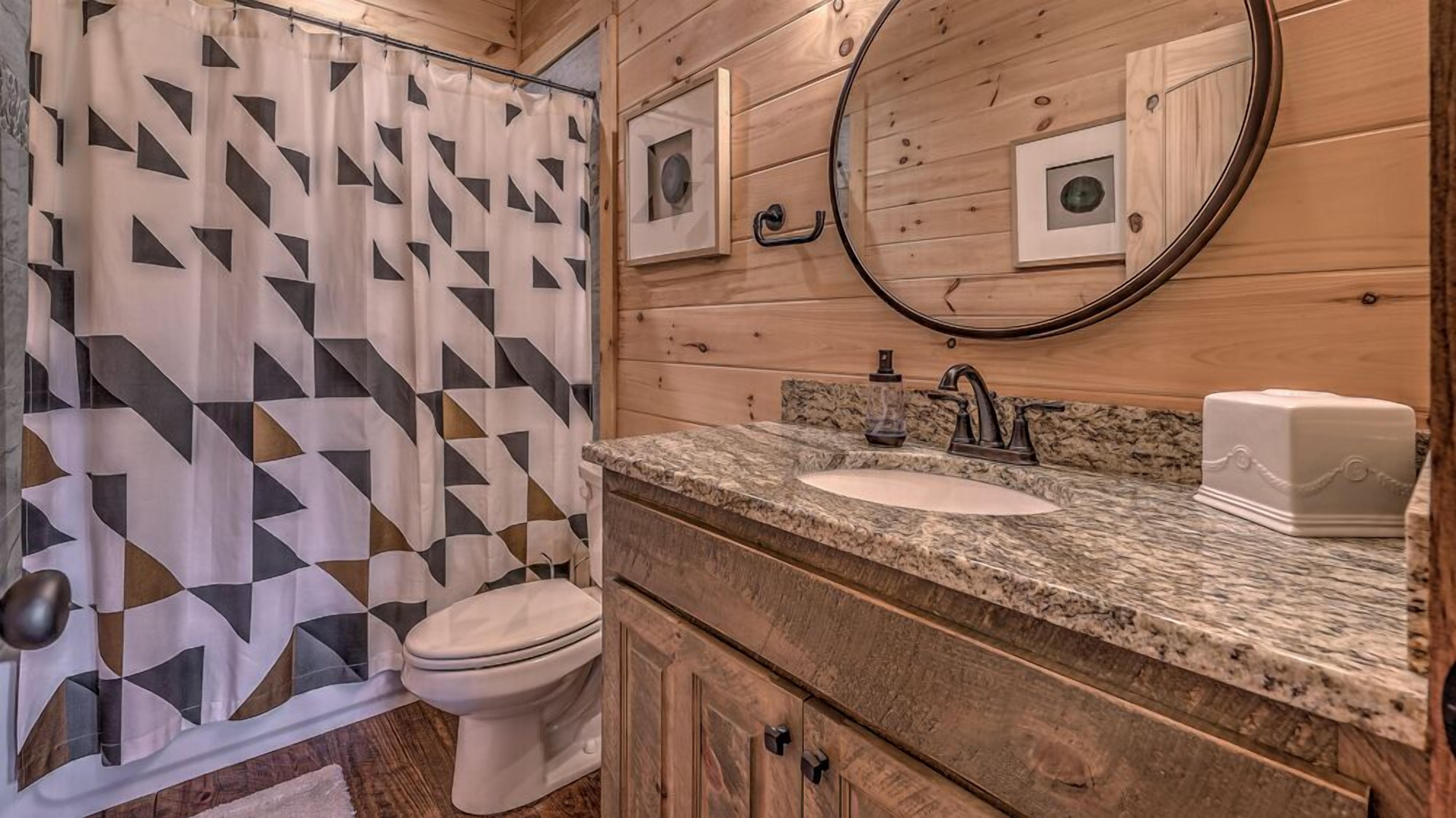 A bathroom with a geometric patterned shower curtain, a white toilet, a granite countertop sink with a faucet, a mirror, and a soap dish.