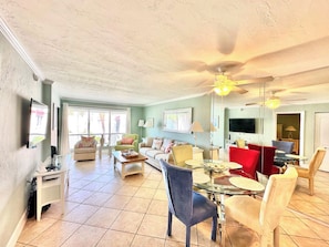 Ein geräumiges Wohnzimmer mit Deckenventilator, einem Essbereich mit Glastisch und einem an der Wand montierten Fernseher.