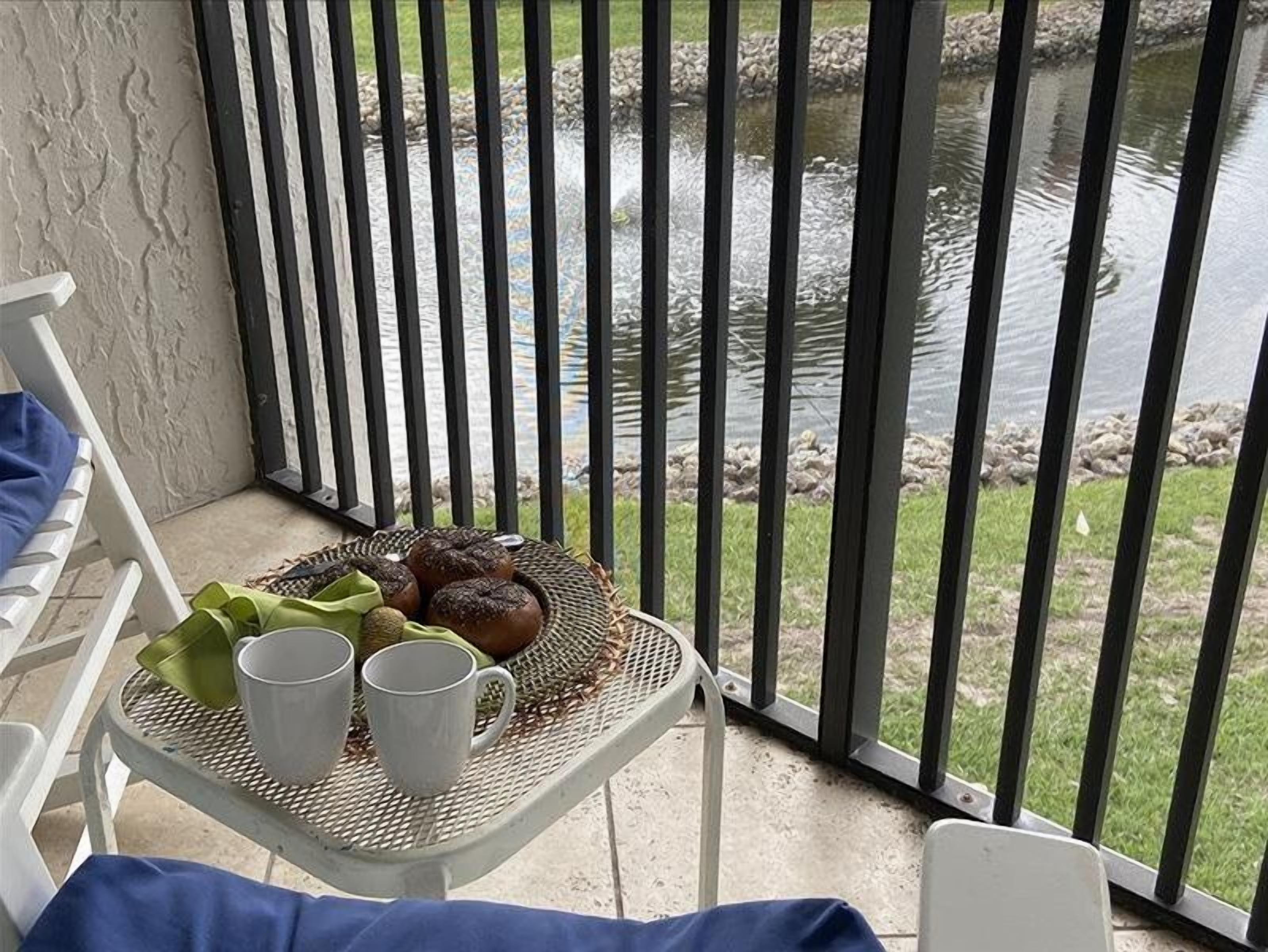 Un balcón con una mesa dispuesta, con pan, un paño verde y tazas blancas, con vistas a un estanque.