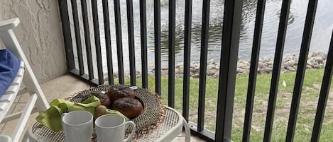 Un balcón con una mesa dispuesta, con pan, un paño verde y tazas blancas, con vistas a un estanque.