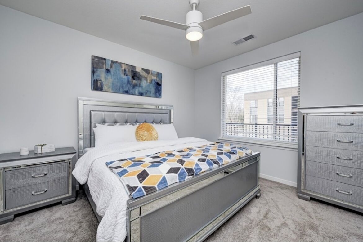 Une chambre à coucher comprenant un lit, une table de chevet, une commode, un ventilateur de plafond et une fenêtre avec des stores.