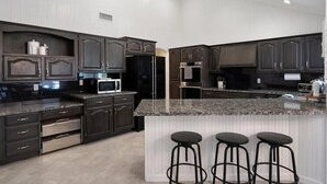 Uma cozinha moderna com armários escuros, bancadas de granito, uma ilha central com banquetas e eletrodomésticos embutidos.