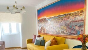 Een moderne woonkamer met een gele bank, een ronde houten tafel en een groot stadsgezichtschilderij aan de muur.