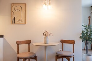 Un comedor con dos sillas de madera, una pequeña mesa redonda, un jarrón con flores secas y un cuadro abstracto moderno en la pared.