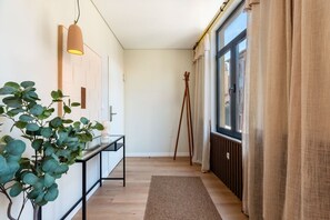 Un pasillo con piso de madera, una consola negra con una planta, una lámpara de pie y una ventana con cortinas.