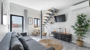 A modern living room with a grey sofa, a staircase, a flat-screen TV, and a large potted plant.