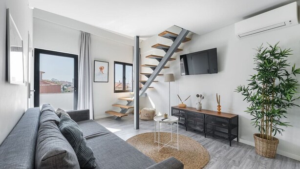 A modern living room with a grey sofa, a staircase, a flat-screen TV, and a large potted plant. - Duplex River View Apartment | With A/C by Lisbeyond (Vila Nova de Gaia)