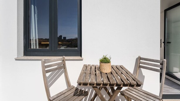 A wooden table with two chairs and a potted plant, positioned next to a window with a view of a cityscape.