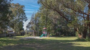 Un parque infantil con tobogán y estructuras para escalar, rodeado de árboles y un césped.