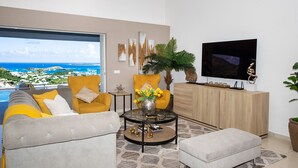 Una sala de estar con sofá, sillón y reposapiés, una mesa de centro con objetos decorativos, un televisor y vista al océano.