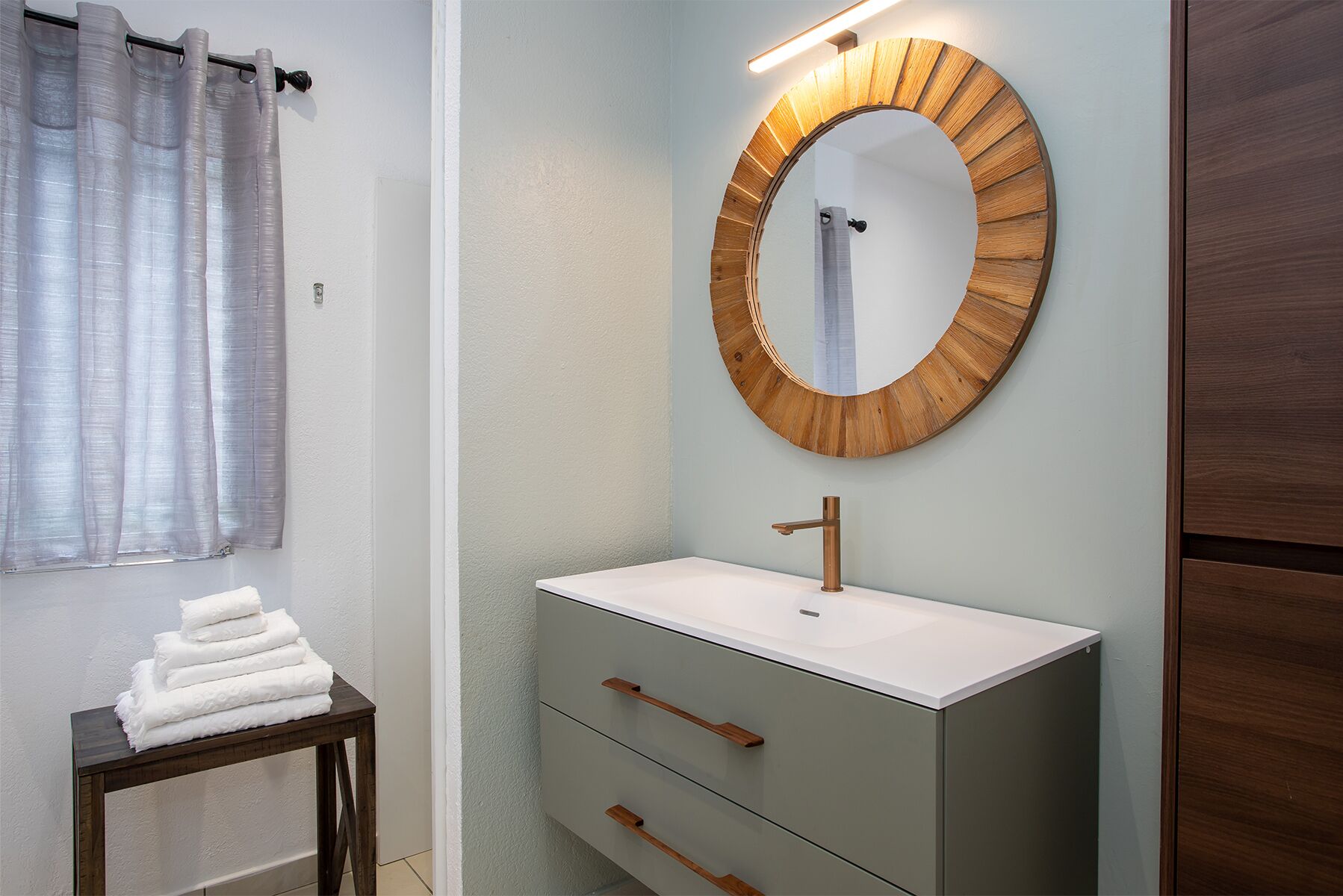Un baño con un espejo redondo, lavamanos blanco y un mueble de madera.