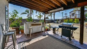 Un patio exterior con zona de bar, taburetes, una parrilla y plantas en macetas.