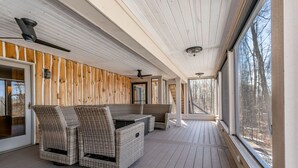 A covered outdoor area with a wooden deck, wicker furniture, and large windows offering a view of the surrounding trees. - Beautiful Rustic Lakehouse in Michigan, gorgeous views and total relaxation (Montgomery)