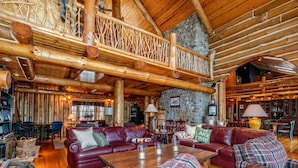 A spacious log cabin interior with a high ceiling, wooden beams, and a stone wall. There are multiple seating areas with leather sofas and a dining table. - Beautiful Rustic Lakehouse in Michigan, gorgeous views and total relaxation (Montgomery)