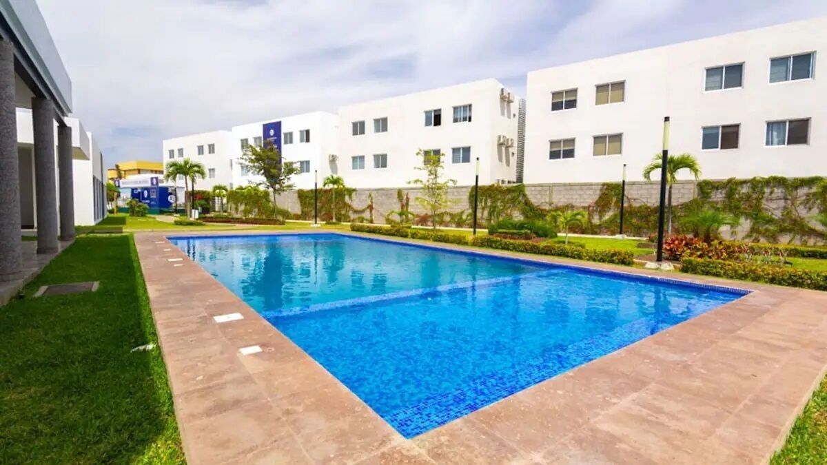 A swimming pool with a tiled edge, surrounded by a well-maintained lawn and a row of white buildings with multiple windows.