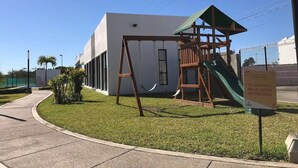A playground with a swing set and slide, a building with a white facade, and a sign in Spanish. - CONDOS AA (Mazatlán)