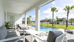 A covered outdoor area with white furniture, a pool, and a view of palm trees and a fence.