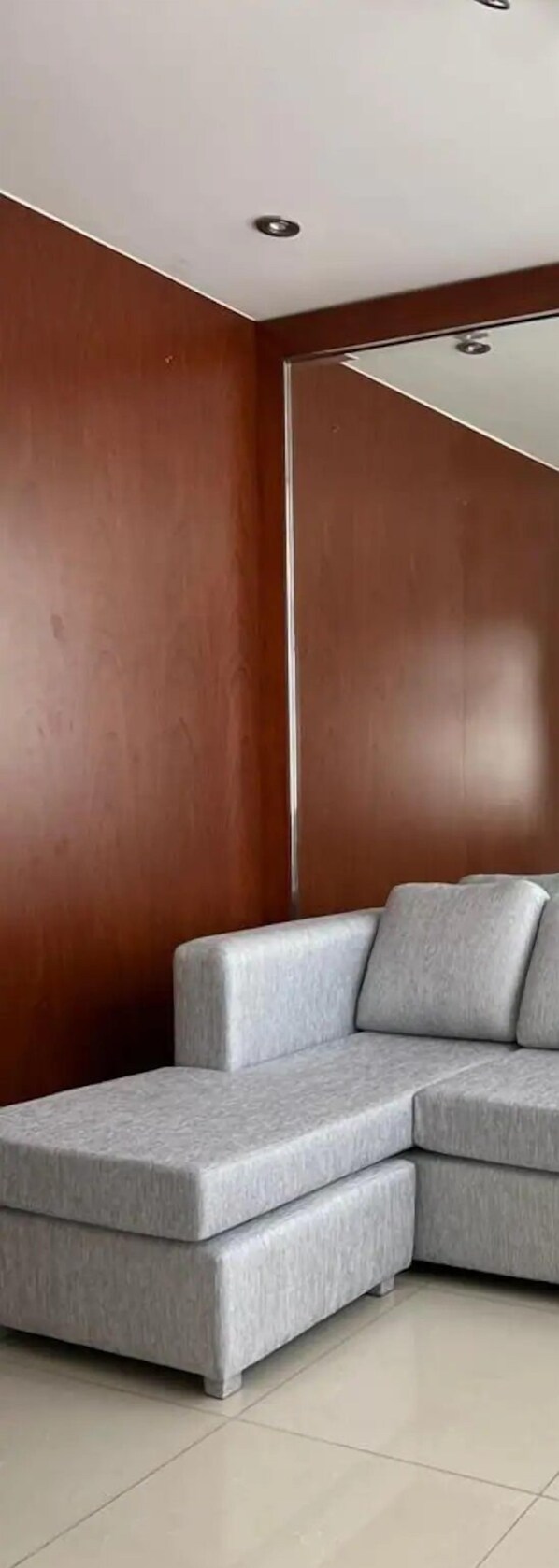 A modern living room with a grey sectional sofa, wooden panel walls, and a recessed ceiling light.