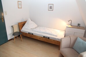 Une petite pièce comprenant un lit simple, une chaise, un petit bureau avec une lampe et un radiateur.