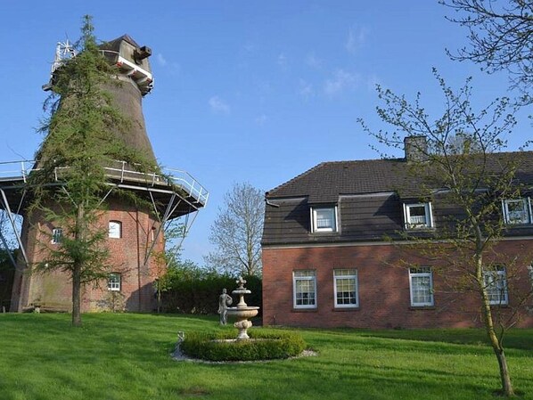 Un moulin à vent traditionnel, avec une base en briques et une structure métallique sur le dessus, situé à c ôté d’un bâtiment en briques à deux étages, doté d’un toit en pente et d’un petit jardin avec une fontaine.