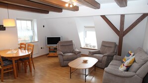 A living room with a wooden dining table, two grey armchairs, a sofa, and a TV on a wooden stand.