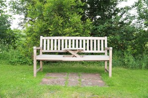 A wooden bench with a backrest and armrests, placed on a stone-paved area surrounded by grass and greenery.