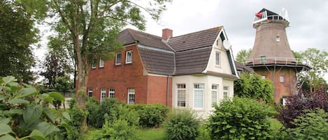 A traditional brick house with a thatched tower, surrounded by lush greenery.