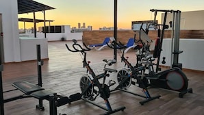 Un gimnasio en la azotea con equipamiento de ejercicio, una banca y vistas del horizonte de la ciudad al atardecer.