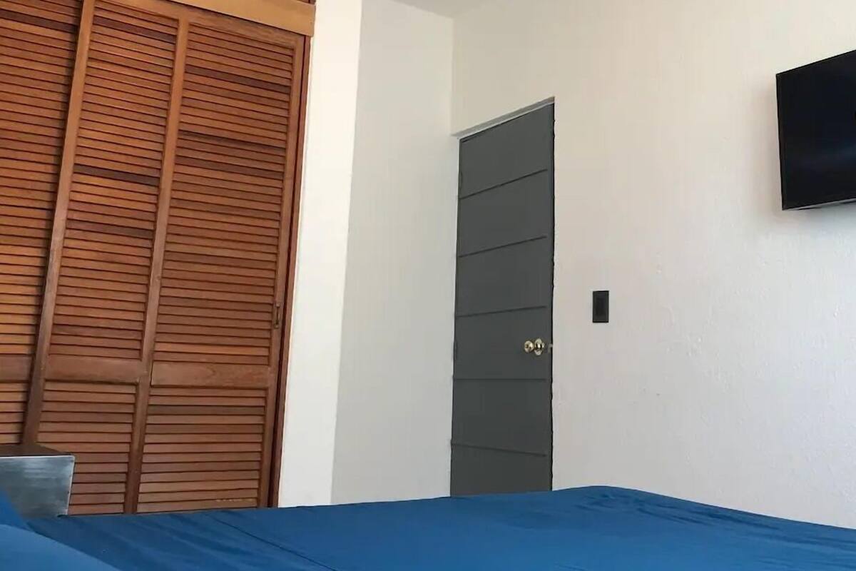 A bedroom with a bed, a wooden wardrobe, a grey door, and a TV mounted on the wall.