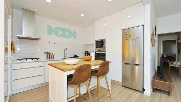 A modern kitchen with white cabinetry, a stainless steel refrigerator, a microwave, and a wooden island with two bar stools. - Tropical Pool View Cascais - Checkinhome (Cascais)